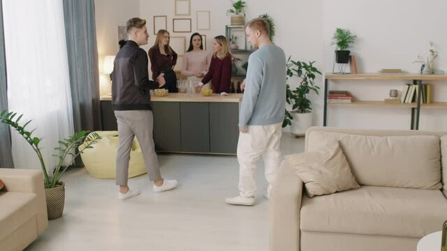 PAN shot of happy young man entering apartment and greeting friend by tapping his foot against his. Group of people having get-together post-pandemic