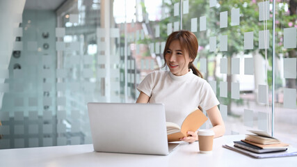 Happy businesswoman sitting near window holding notebook and using computer laptop.