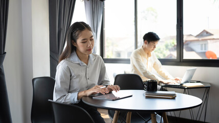 Fototapeta premium Young asian office worker using digital in office and her colleague sit-in in background.