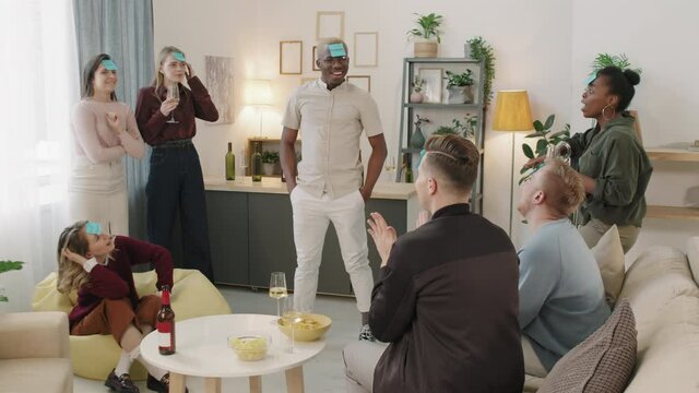 Medium shot of group of happy young men and women with sticky notes on their foreheads having fun at get-together in cozy apartment. They are drinking alcohol and playing guessing game