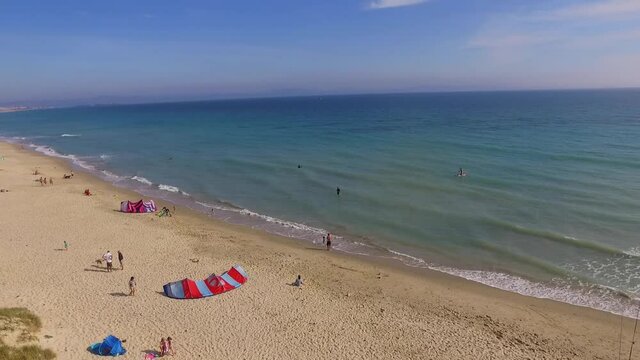 Paning view of the coast. Waves crashing on the beach line, some fishing rods, a couple unmbrellas and a kite surf seen on the sand.