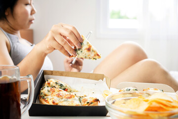 lazy Asian woman eating junk, fast food in bed hand holding pizza with bowl of potato chips and glass of sweet soda on table