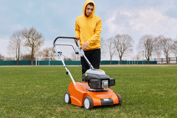 Handsome young adult male worker mowning field with grass cutter, guy looking at lawnmower and tries to turn on, man wearing yellow hoodie and black trousers cutting grass.