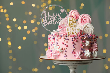 Beautiful birthday cake with decor on stand against festive lights. Space for text