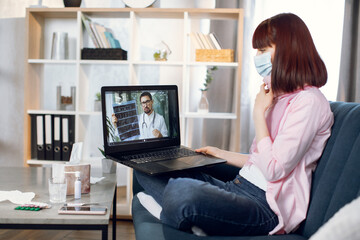Young woman covered with thick blanket, sitting on a bed in a bright room and having an on-line consultation with her doctor