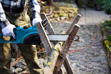 Man with electric chain saw while cutting firewood. Seasonal working in the garden. Worker working...