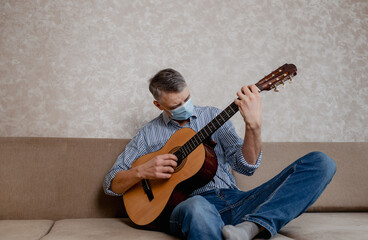 a man in a medical mask plays guitar at home on the couch. Home entertainment in isolation