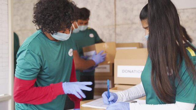 Two Volunteers Discussing About Donated Stock, Checking And Writing On Clip Board While Other Busy Arranging Donation Boxes And All In Medical Face Mask And Gloves As Covid-19 Pandemic Safety Measures