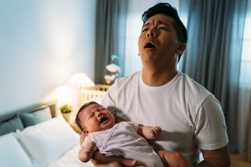 portrait overwhelmed asian father is holding his crying baby daughter and looking upward in despair at midnight in the bedroom at home.