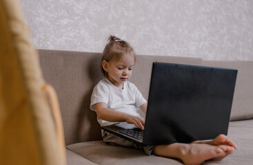 A little girl is sitting on the couch at home with a laptop. A girl looks at a computer screen during online classes with a happy smile