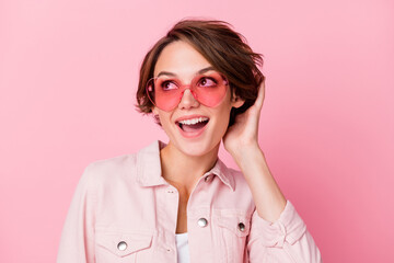 Photo portrait of cute girl touching hair in rose-tinted heart-shaped glasses isolated on pastel pink colored background