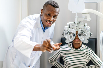 Optician doing optometry eye exam for black african american teen girl patient. Male optometrist with phoropter while examining patient. - obrazy, fototapety, plakaty