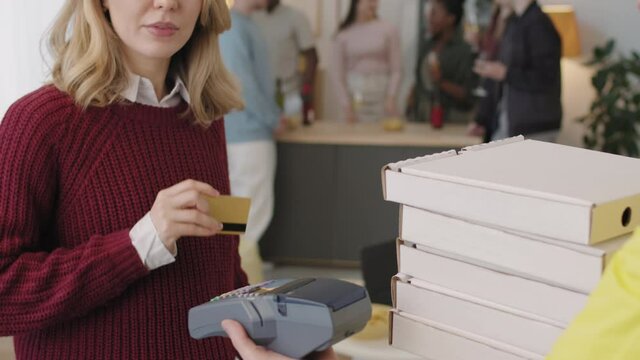 Slowmo of happy young woman touching debit card to terminal held by deliveryman and paying for pizza in her apartment. Group of friends chatting in background