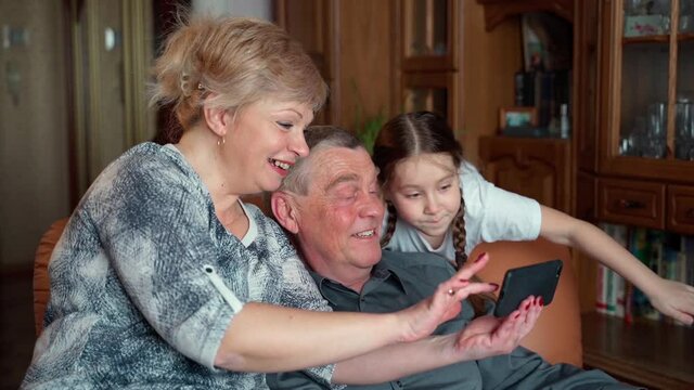 Slow motion shot grandparents selfie with granddaughter at home. Senior grandpa and grandma happy spend family time relax using mobile phone technologies with young girl sitting on sofa in living room