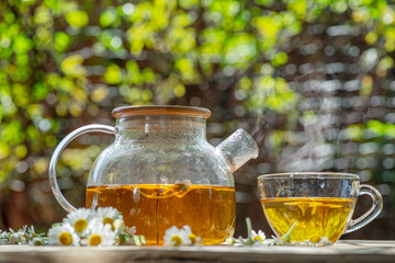 Herbal chamomile tea and chamomile flowers near teapot and tea glass on wooden table. Countryside background.