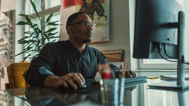 Handsome Black Male Specialist Sitting at His Desk Works on Computer in Creative Office. Professional works on Mobile Software Development, App Design, Social Media Project. Side Medium Portrait
