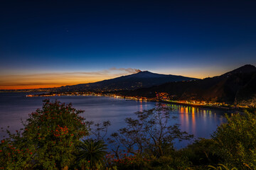 Fire and lava flowing on the top  of mount Etna during eruption. Night scenery on the Mediterranean coast, city lights
