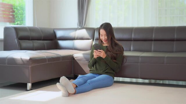 Asian Woman Sitting on the Floor near Sofa Texting on Smartphone.