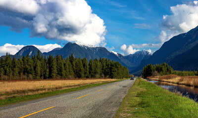 A country road in the Pitt River Valley runs through farm fields and forest to  Pitt Lake on the background of the snow-covered mountain range and a beautiful cloudy sky