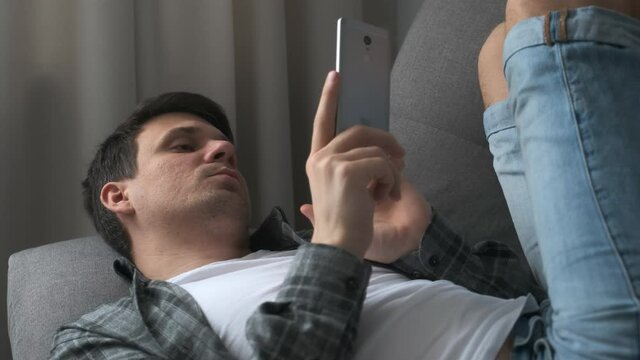 Young man is browsing smartphone lying on sofa finding a woman on dating site online. He is showing no and yes gestures and smiling. Virtual life of millennial man. Swipes pages on phone.