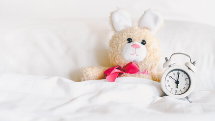 analog alarm clock and blurred cute little rabbit doll lying alone on white bed for wake up or go...
