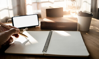 Focus on human hand holding pen write on book and using mobile phone meeting online, work at home on wooden table near bright window with sunlight shining through, vintage tone