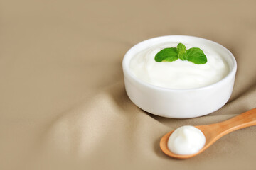 Yogurt natural flavor in white ceramic bowl with peppermint leaf and wooden spoon placed on light brown cloth with copy space, Milk curd organic homemade for health care and healthy food concept.