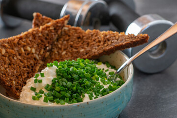 Workout meal with low fat cottage cheese, chives and wholegrain bread with dumbbells in the background - obrazy, fototapety, plakaty