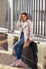 Caucasian portrait of a lovely brunette girl in a light coat and blue jeans with a smile stands near the railing on a sunny spring day. The model happily shows his hands to the sides.