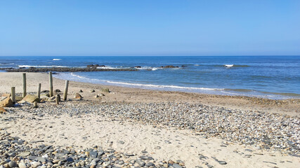 Sea, beach boulders, pebble shore and waves. Sea waves breaking on rocks. Sunny beach with sand dunes and blue sky in Cepaes, Esposende, Portugal.