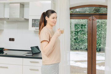 Young woman in casual clothes is drinking cappuccino or caffee in her kitchen. Housewife is happy in the kitchen in the morning 