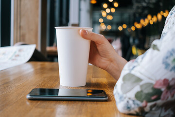 White large paper cup of tea on a table in a cafe. A woman's hand holds the cup, a smartphone lies...