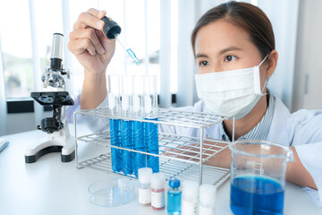 Scientist woman in medical face mask is holding dropper to dropping chemical liquid into test tube