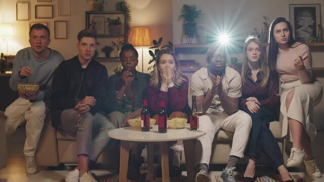 Medium shot of multi-ethnic group of young people sitting on couch and watching suspenseful movie, then getting frightened by jump scare