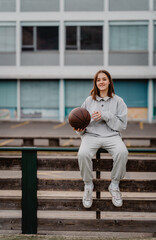 Teenage girl holding basketball ball sitting on sportground fence smiling. Teen lifestyle, sport, outdoors leisure activity.