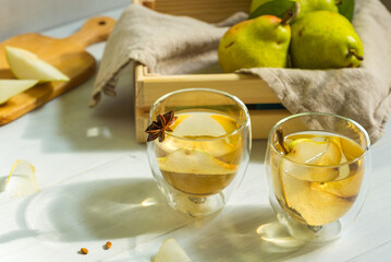 Two glasses of refreshing pear cider on a wooden table, lit by the sun from the window. A small box with pears in the background