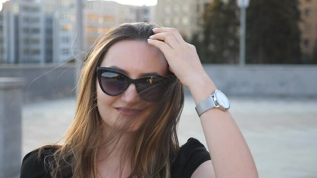 Portrait of young businesswoman in sunglasses with cityscape at background. Happy attractive business woman looking at camera and straightens hair. Face of smiling confident girl. Slow motion Close up