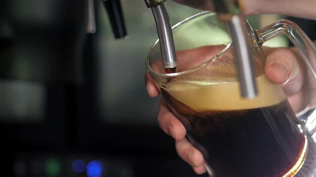 The bartender pours a dark beer into a beer glass behind the bar. The bartender pours craft beer at the bar. Close-up