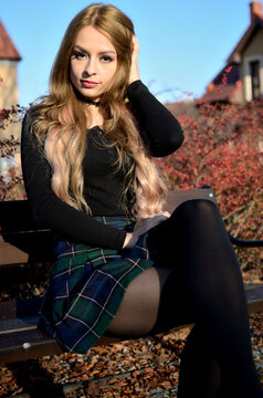 Charming young woman with blonde hair extension sitting on bank in small town. Polish model with black tights, short skirt and black top.