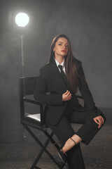 Pretty caucasian woman with long straight brunette hair sitting at high chair with crossed legs. Fashion female woman wearing black male style suit with tie