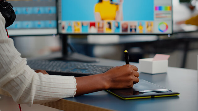 Close up of african photo retoucher holding stylus pen editing customer photo using computer with two displays. Graphic editor retouching assets drawing on graphic tablet in creative agency office