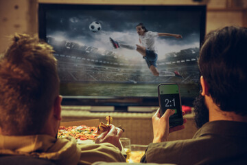 Men with betting application in phone. Group of friends watching TV, sport match together. Emotional fans cheering for favourite team, watching on exciting game.