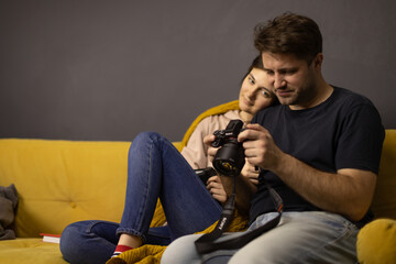 Attractive millennial couple sitting on comfy yellow sofa looking at photos on DSLR screen copy space. Girlfriend and boyfriend reviewing video on screen of digital camera. Family Hobby Concept
