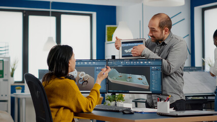 Team of architects and engineers working on building prototype project, using computers running 3D CAD Software. Project manager discussing with employee holding tablet explaining technical specifics - obrazy, fototapety, plakaty