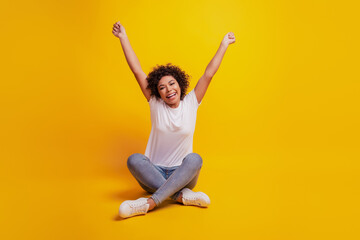 Portrait of beautiful young woman celebrate victory raise hands crossed legs on yellow background