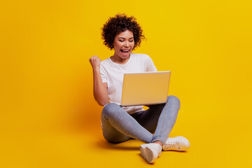 Smiling woman typing on laptop computer raise fist scream open mouth on yellow background