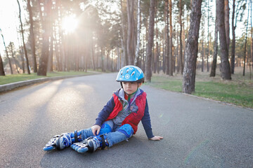 Sad kid in sport helmet sitting on road during riding on roller skates at park, active family weekend, spring outdoors activity, healthy lifestyle