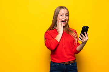Fototapeta premium Shocked woman in sweater holding smartphone and rejoices over yellow background