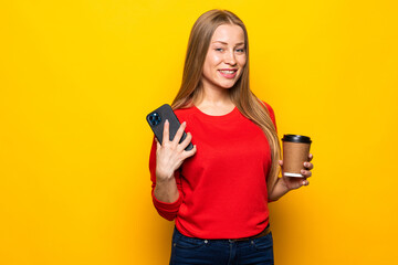 Beautiful young woman drinking coffee using mobile phone posing isolated over yellow wall background