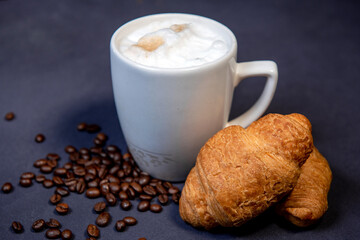 Beautiful latte macchiato with milk foam in a white cup with croissants and coffee beans nearby. Dark blue background. A cup of coffee with milk. Hot latte or cappuccino on milk with copy space